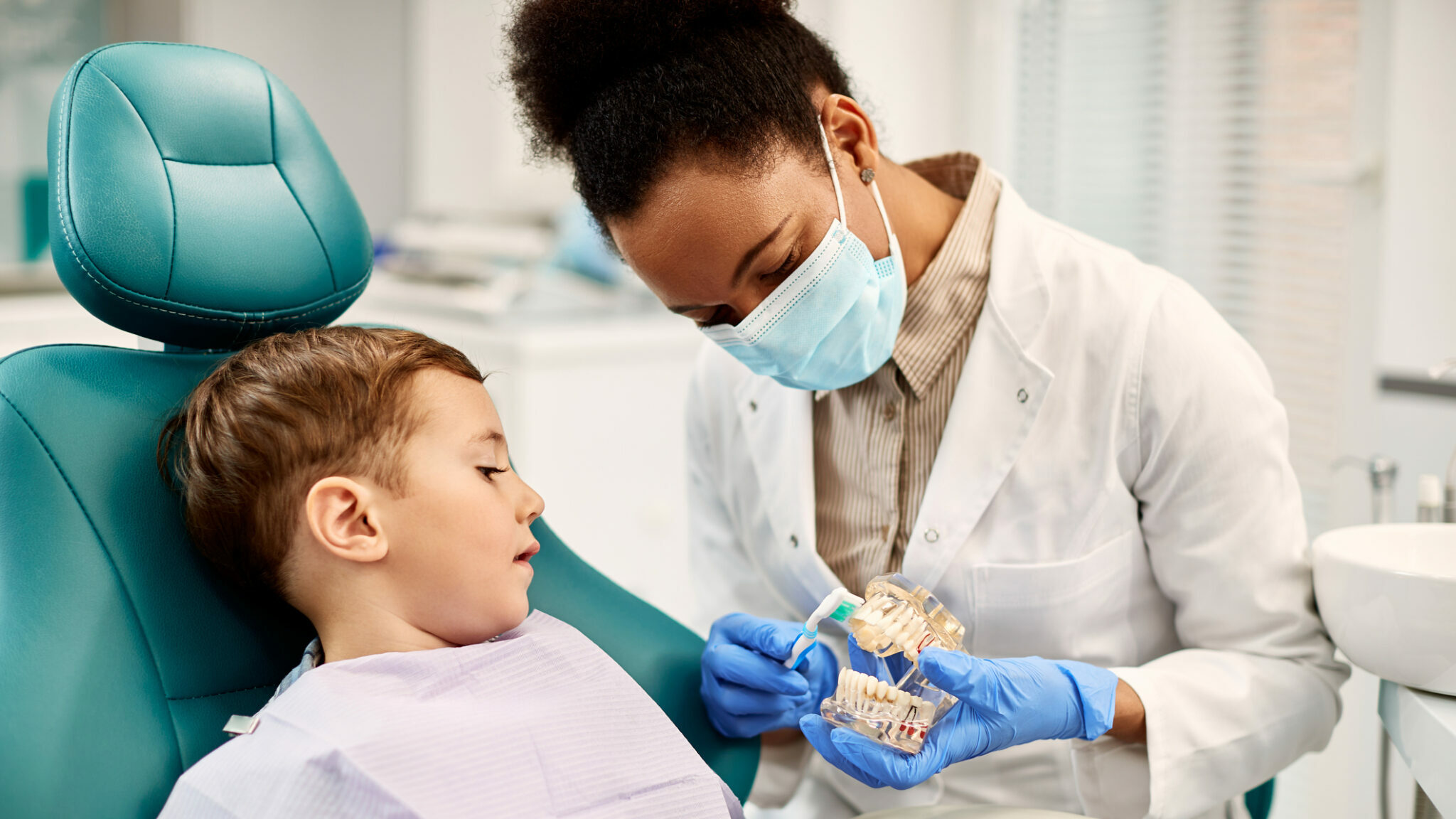 African American dentist teaching small boy how to brush teeth properly during dental appointment. alternate text for this image