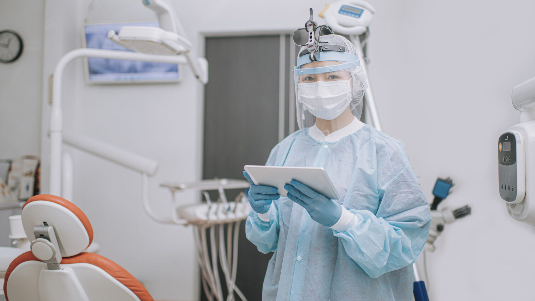 asian chinese dentist with PPE protective workwear holding digital tablet in dentist’s office looking at camera alternate text for this image