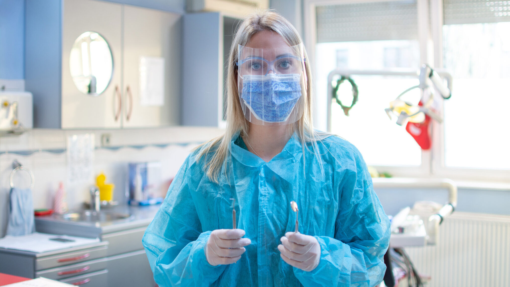 Female dentist in a protective suit with a protective shield and mask holding dental tools in dental office. alternate text for this image