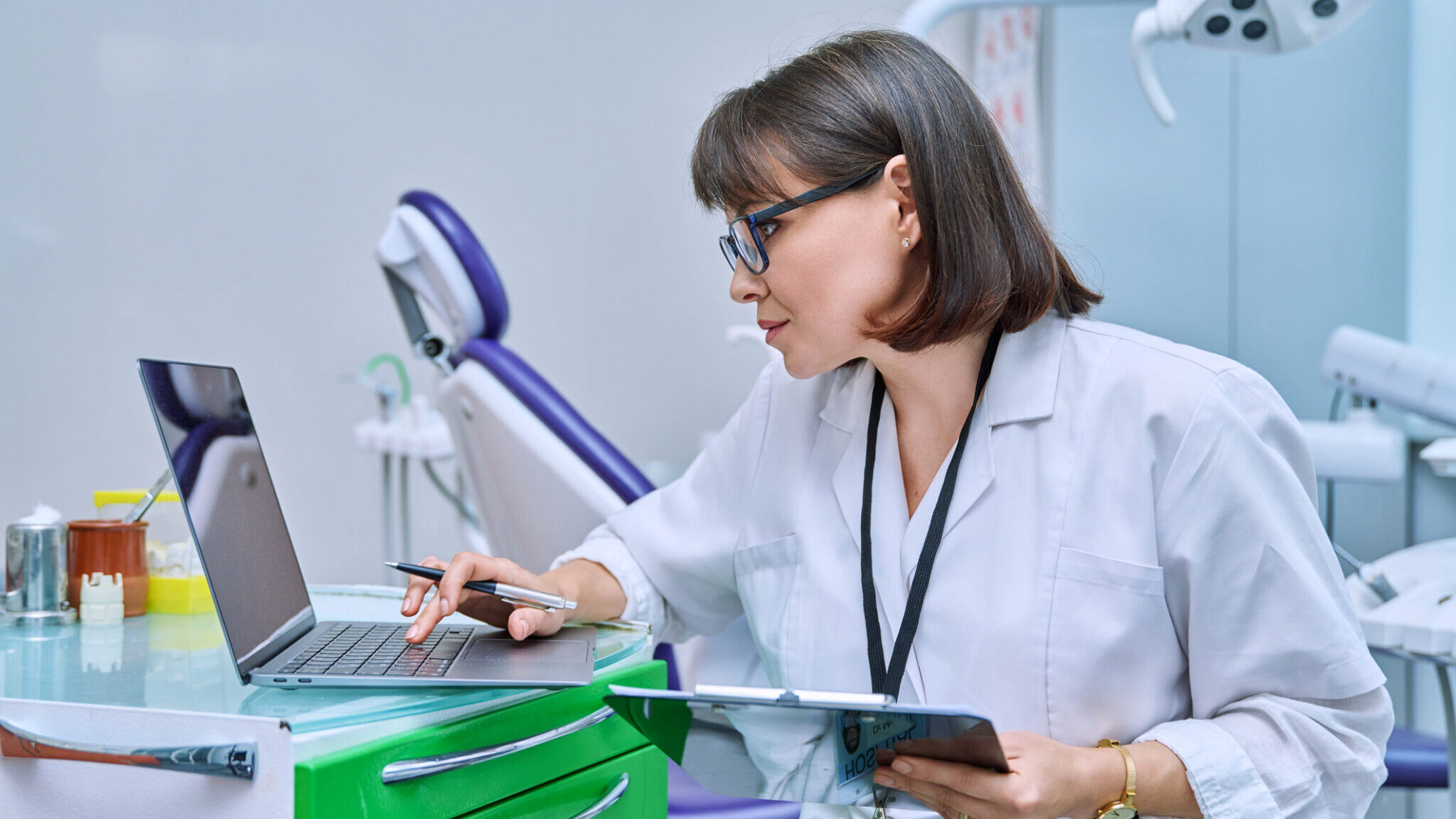 Doctor dentist working in office, using laptop, making notes on clipboard alternate text for this image