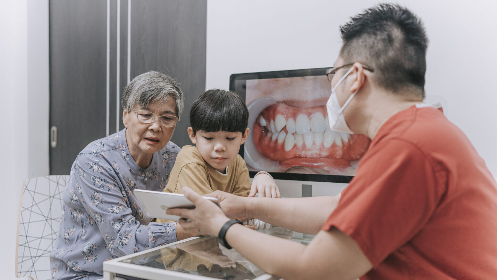 Asian Chinese male dentist explaining to senior woman and young boy on teeth structure and discussion on tooth whitening repairing with monitor screen teeth image alternate text for this image
