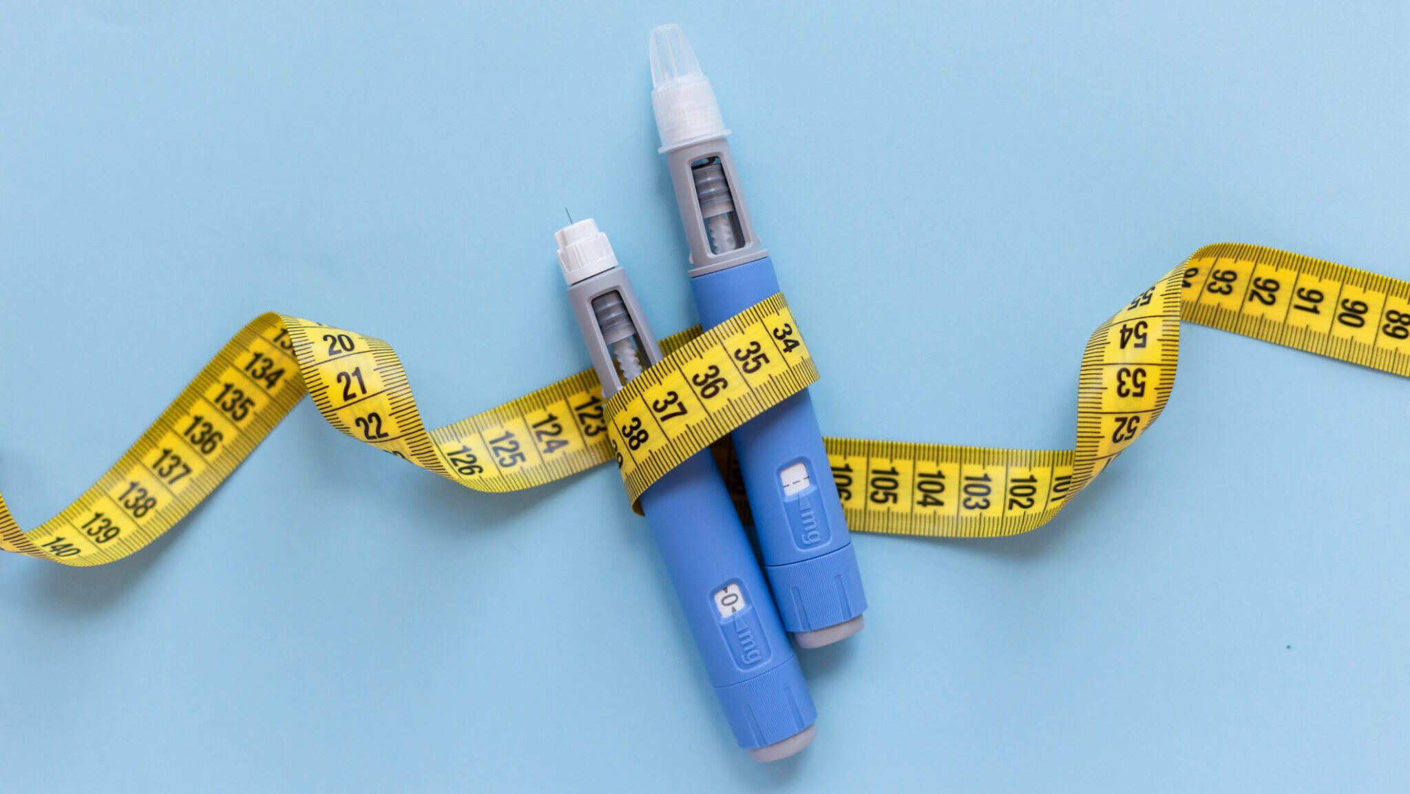 Two injectors dosing pens for subcutaneous injection of antidiabetic medication or anti-obesity medication hovering over a blue background. Yellow measuring tape around the injectors.