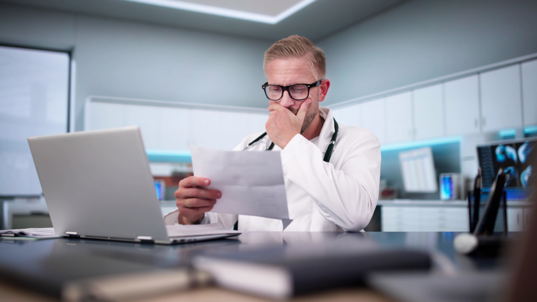 Shocked Medical Doctor Reading Document At Desk In Office