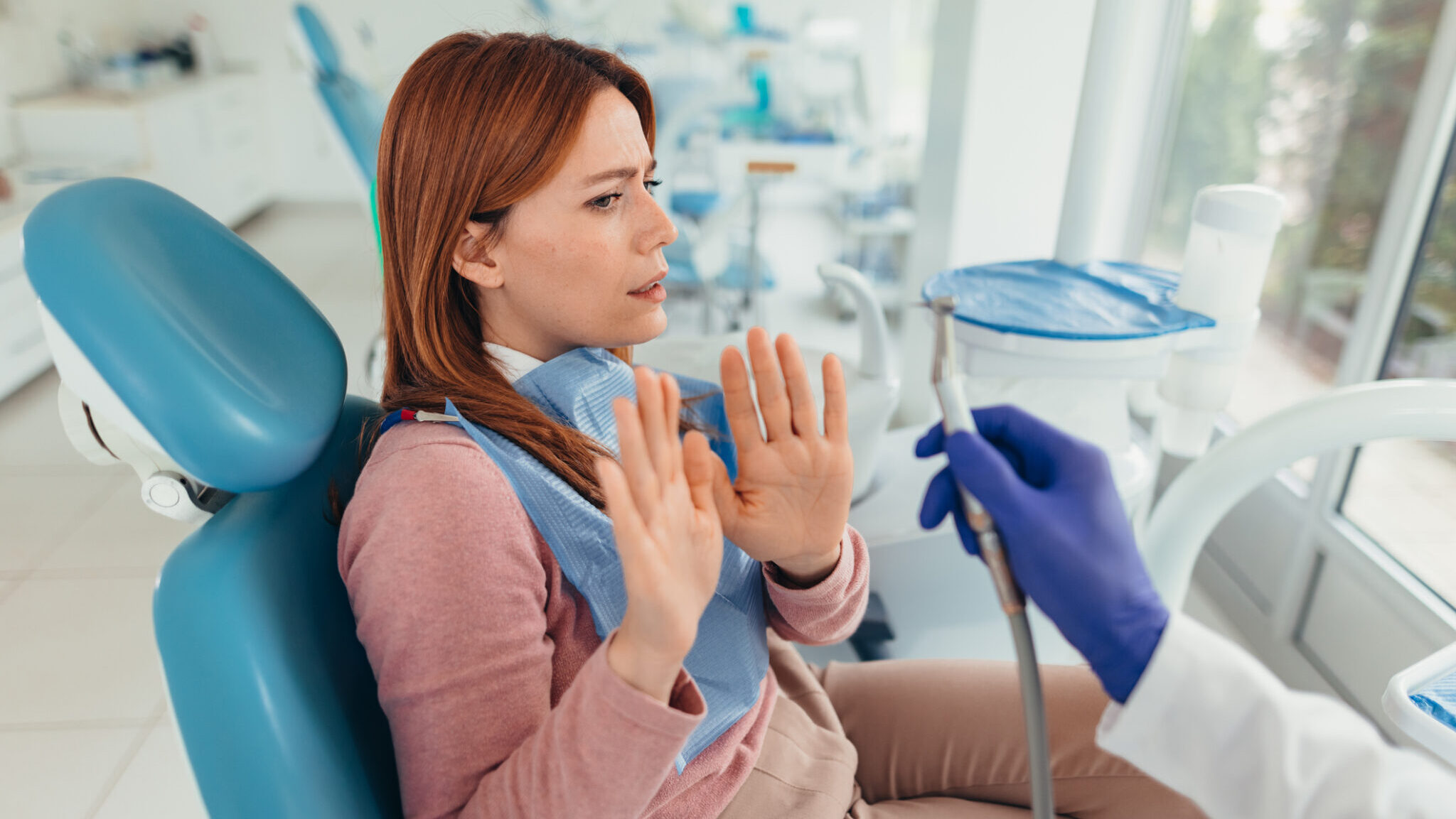Scared patient showing stop gesture with hands sitting on dentist chair refusing dental treatment while dentist holding drill approaching her