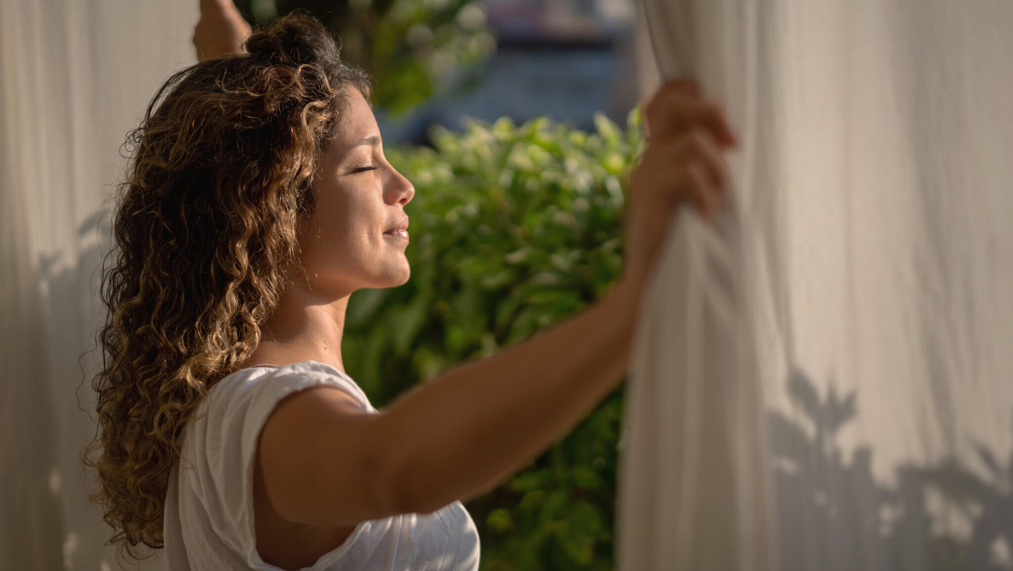 Portrait of a peaceful woman looking through her bedroom window in the morning and felling the sun in her face