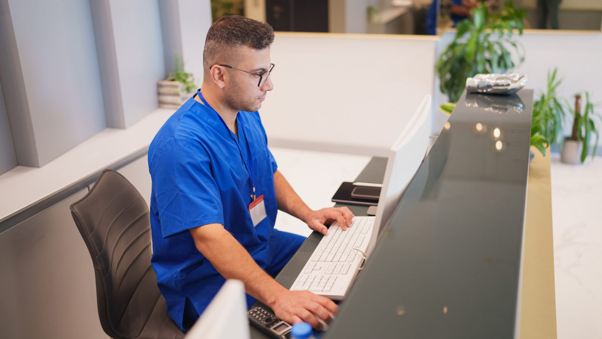 Dental clinic front desk receptionist in blue medical scrubs working on a computer, managing patient records and appointments in a modern, clean healthcare environment.