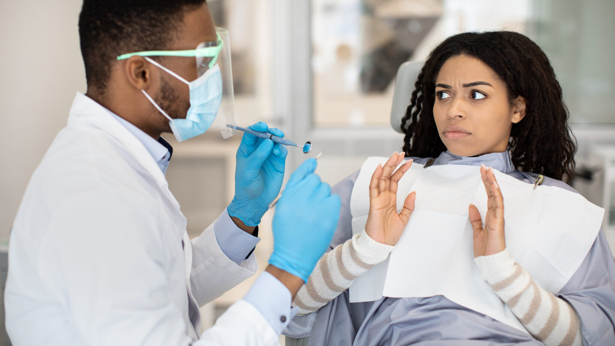 Worried Black Female Patient Looking At Dentist Doctor With Shock During Check Up In Clinic, Scared African American Woman Suffering From Dental Treatment Phobia, Showing Stop Gesture With Hands