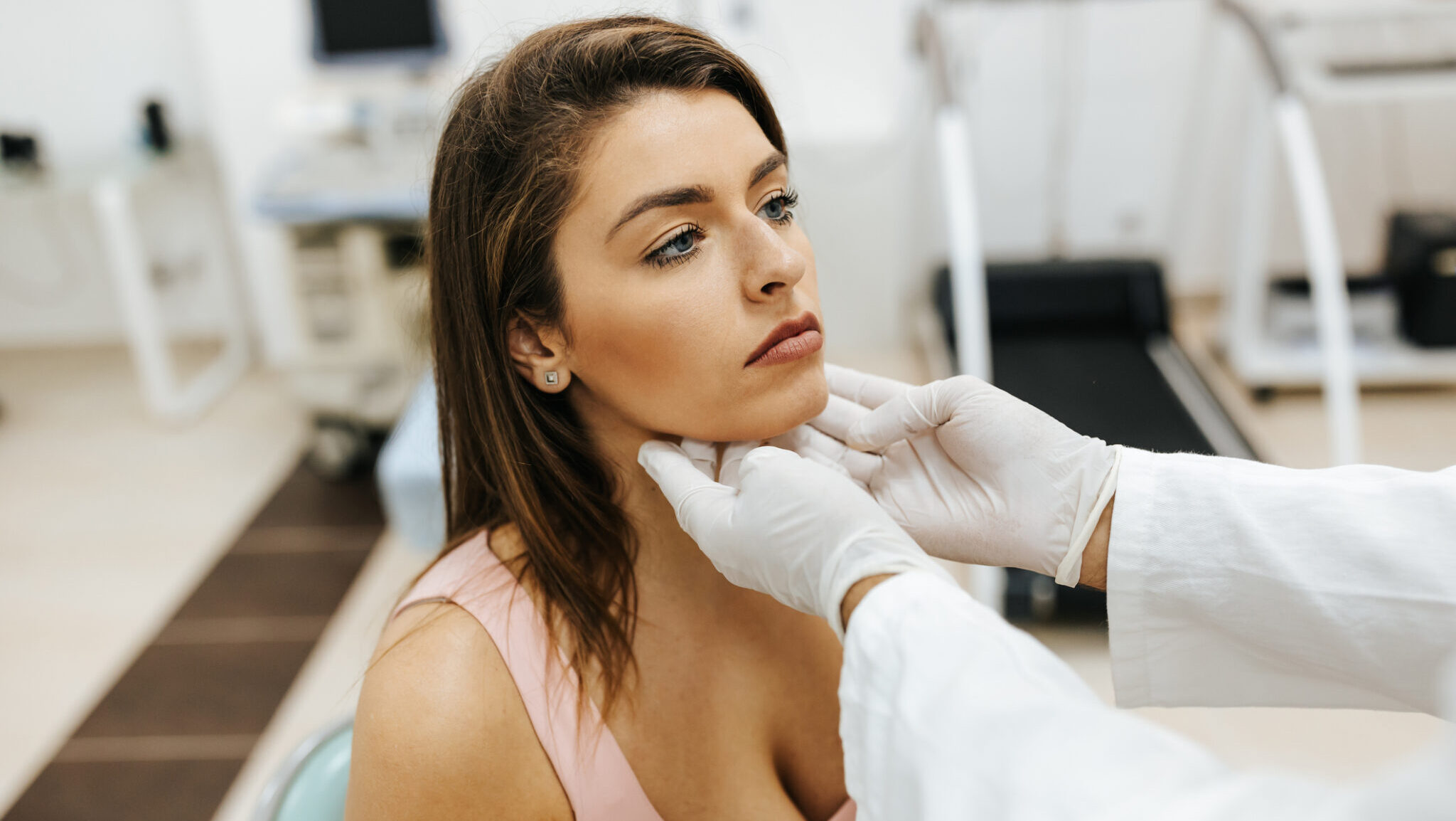 Close-up of young woman visiting general practitioner due to soreness and pain in her throat. The doctor is touching her neck to see if there are any swollen glands